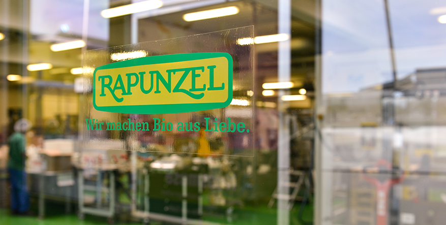 Blick durch eine Scheibe in eine Poduktionshalle. Auf der Scheibe ist ein Aufdruck des Rapunzel-Logos.
